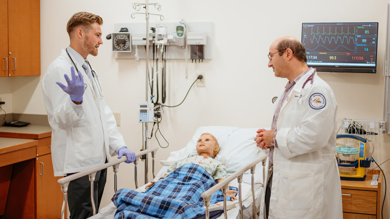 A student and a faculty member, both in lab coats, assess a pediatric manikin in a hospital bed.